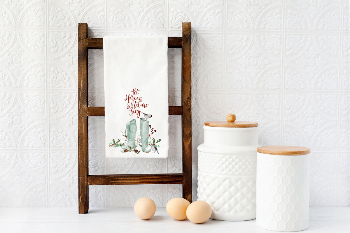 A white kitchen towel with a 'Merry & Bright' holiday design hanging on a dark wooden ladder rack, styled with white ceramic canisters and three eggs against a white textured tile wall.