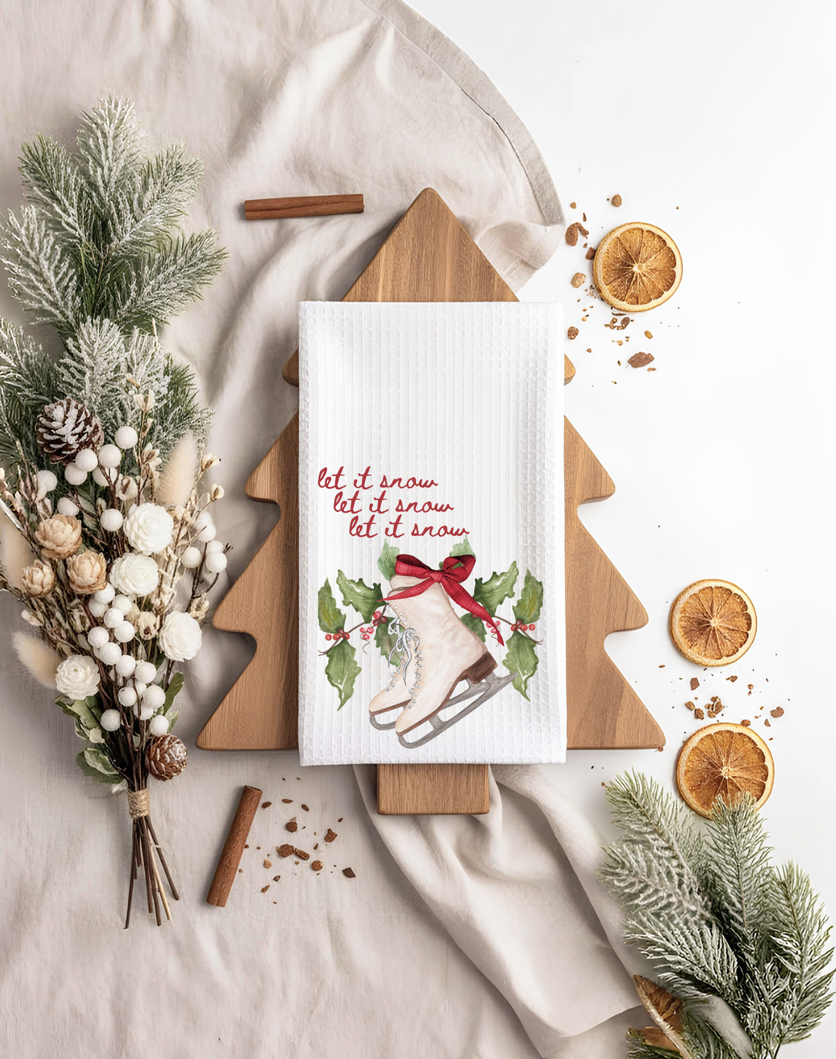 A white waffle-weave kitchen towel with a Christmas theme, featuring the text 'let it snow let it snow' and an illustration of a boot in the snow with holly and a red bow. The towel is displayed on a wooden tree-shaped board, surrounded by holiday decorations including pine branches, dried orange slices, cinnamon sticks, and a dried floral arrangement on a beige fabric background.