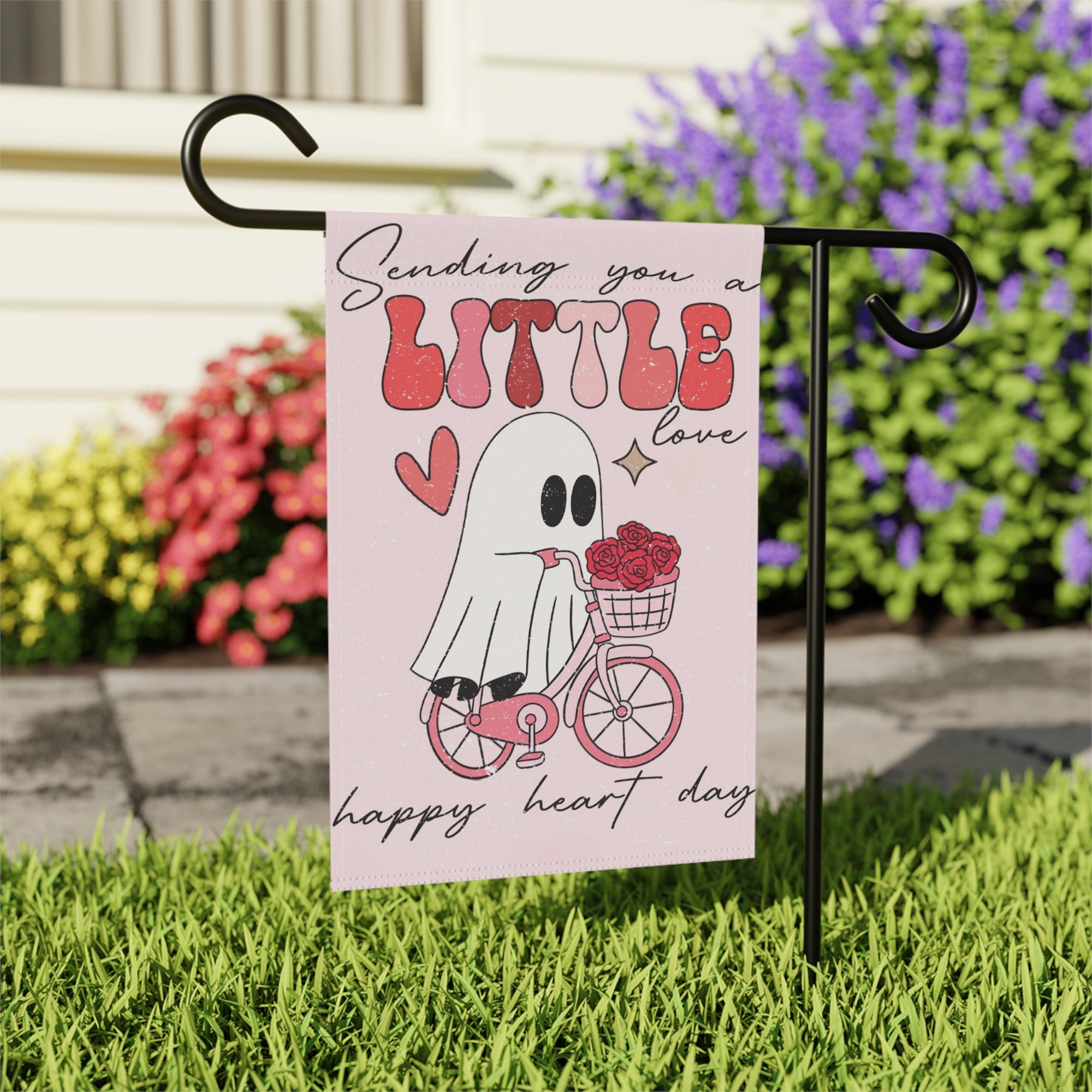 Decorative garden flag with a ghost on a bike and text 'Sending you a little love happy heart day' in a garden setting.