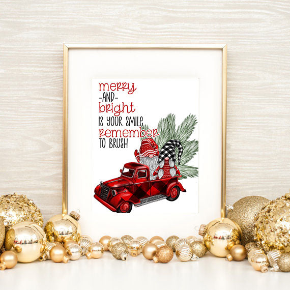 Framed Christmas-themed print with a red truck and gnome, surrounded by gold ornaments on a light wooden surface.